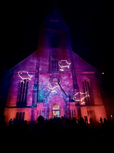 Farbenfrohe Lichtprojektionen auf einer Kirche bei Nacht, Menschen versammelt davor.