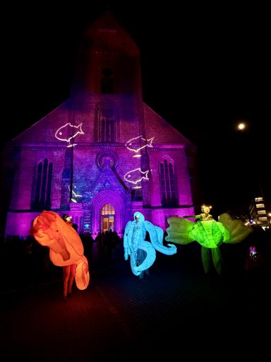 Bunte Lichtinstallationen und tanzende Figuren vor einer beleuchteten Kirche nachts.