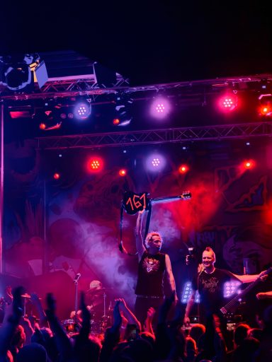 Rockband spielt live auf einer Bühne mit bunten Lichtern und jubelndem Publikum.